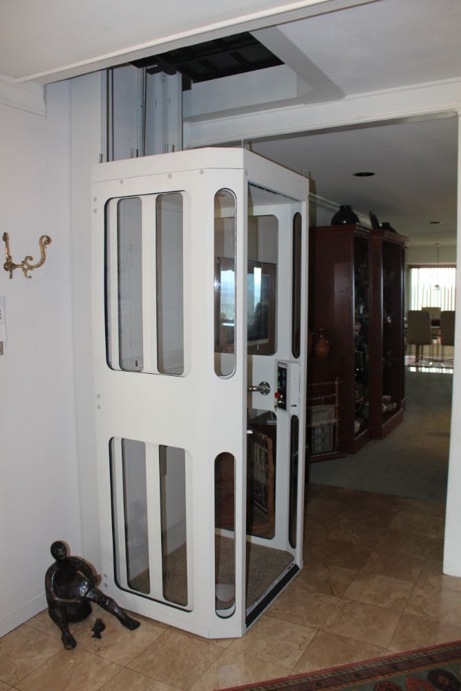 A white residential floor-to-floor elevator standing in a tiled home interior, positioned under an open ceiling cutout.