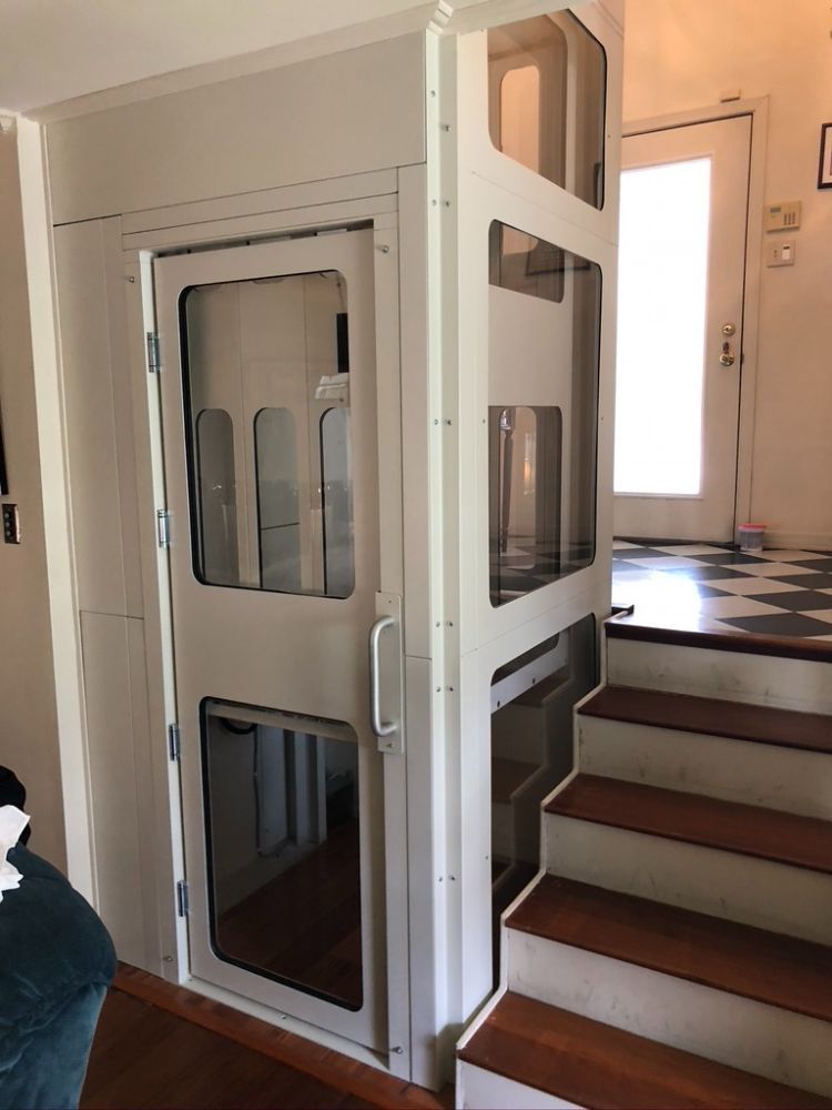 A white, enclosed residential elevator installed next to a wooden staircase inside a home.