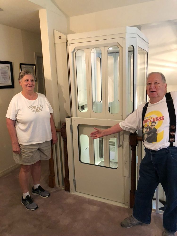 Two people stand in a home next to an indoor residential elevator with a white frame and glass door.
