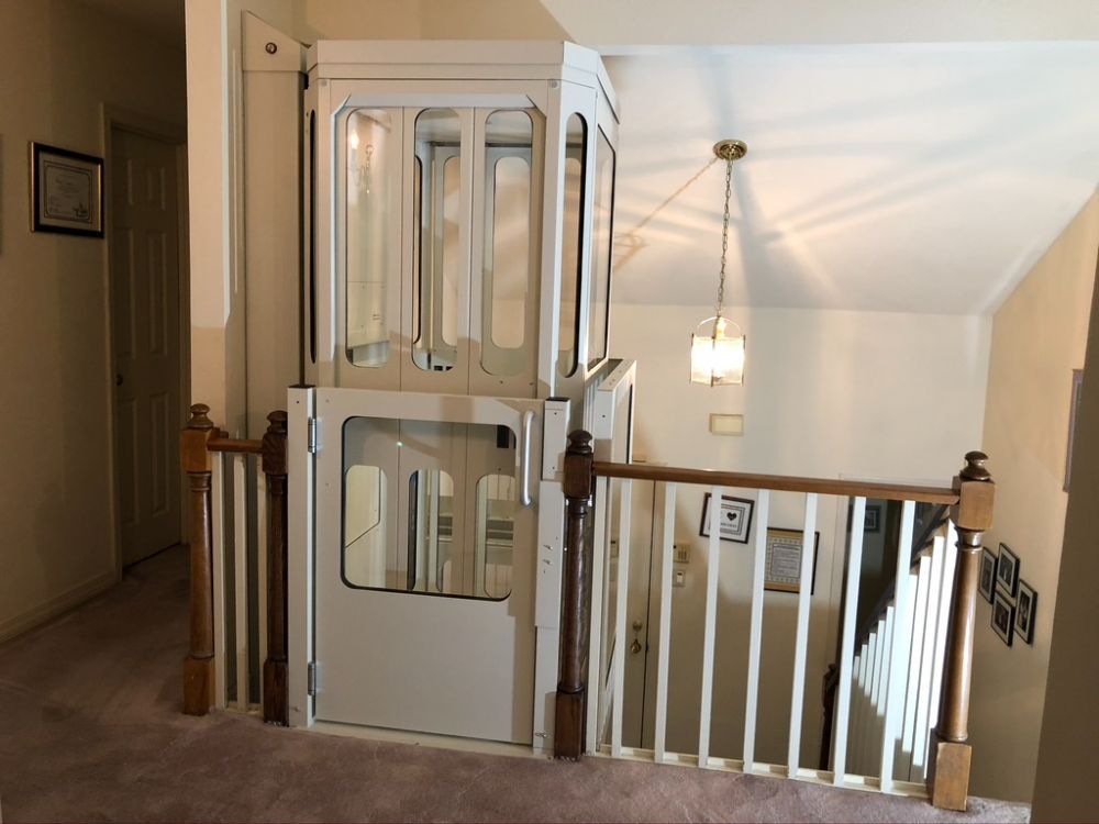 A white residential home elevator with glass panels installed next to a wooden staircase railing on an upper floor.