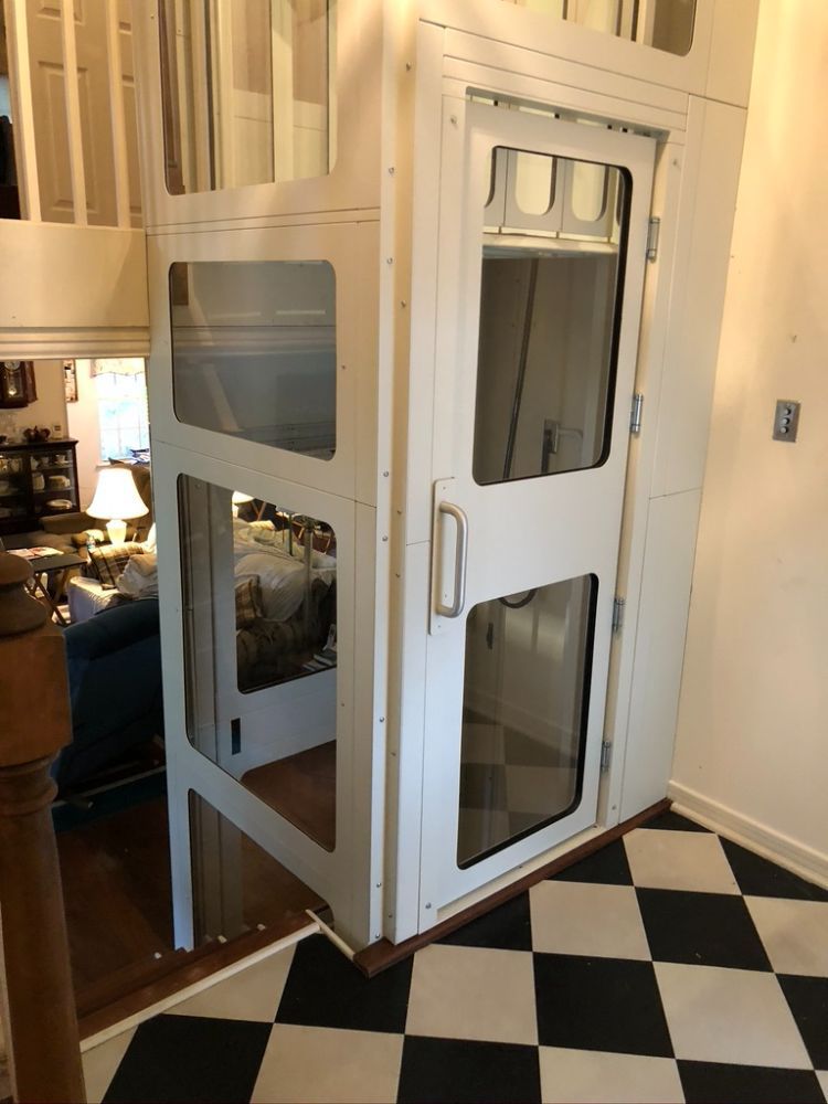A white, framed residential elevator with glass panels stands on a black-and-white checkered floor in a home.