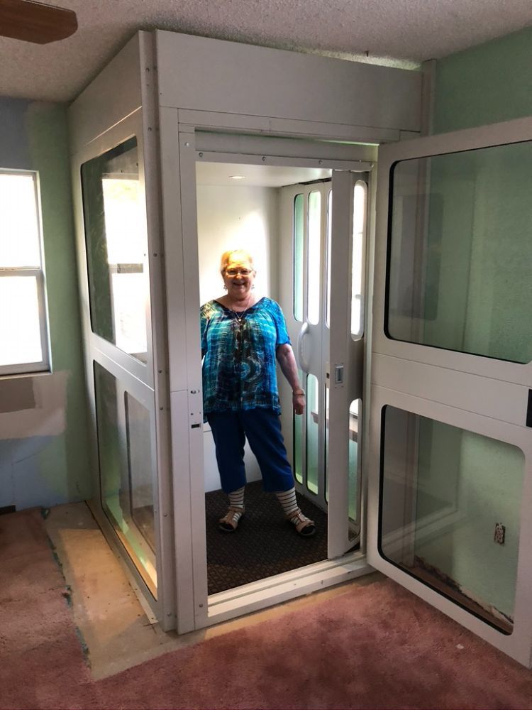 A person stands inside a white residential elevator unit installed in a room with light green walls and pink carpet.