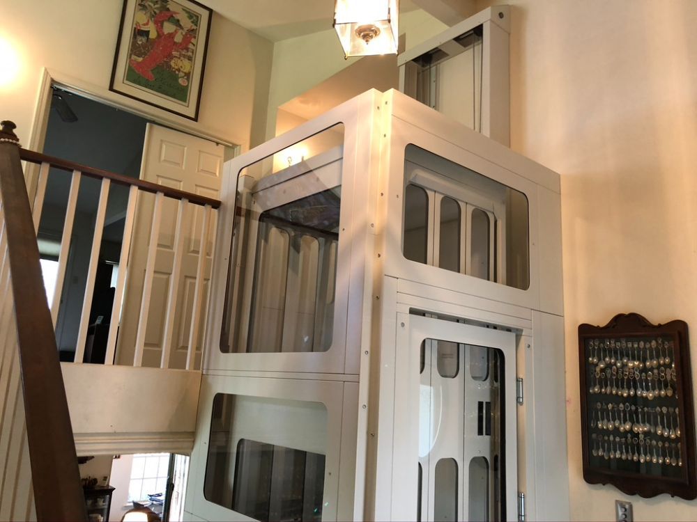 A white, enclosed home elevator with glass panels stands next to a wooden staircase inside a house.