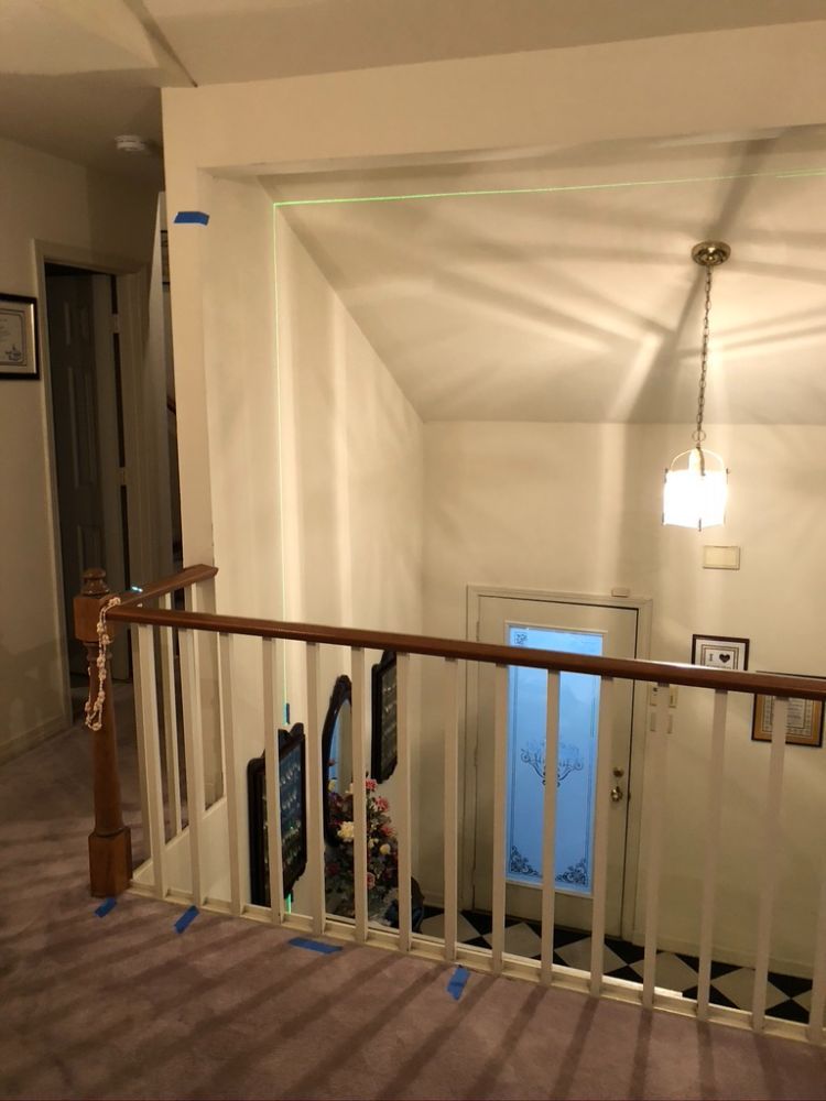 A laser level line stretches across a white wall above a staircase, with blue painter's tape marked along the banister.