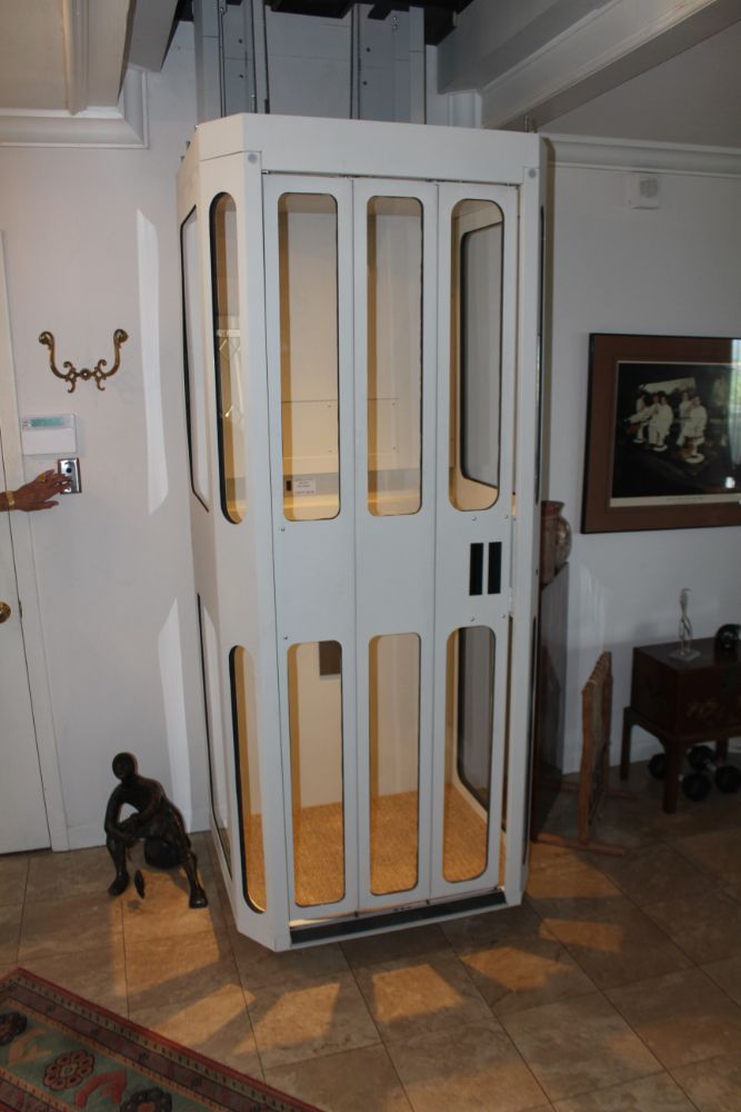 A white, glass-paneled residential elevator stands in a tiled room with a person’s hand operating the call button.