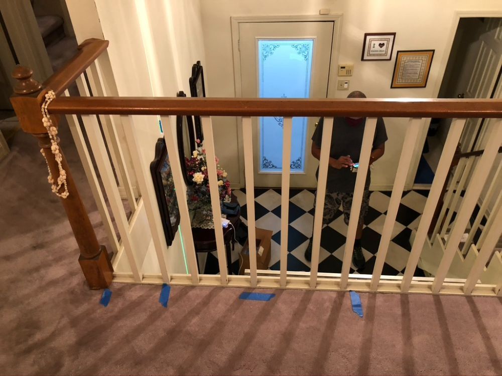 A balcony railing with small blue adhesive strips on the floor, overlooking an entryway with checkered flooring.