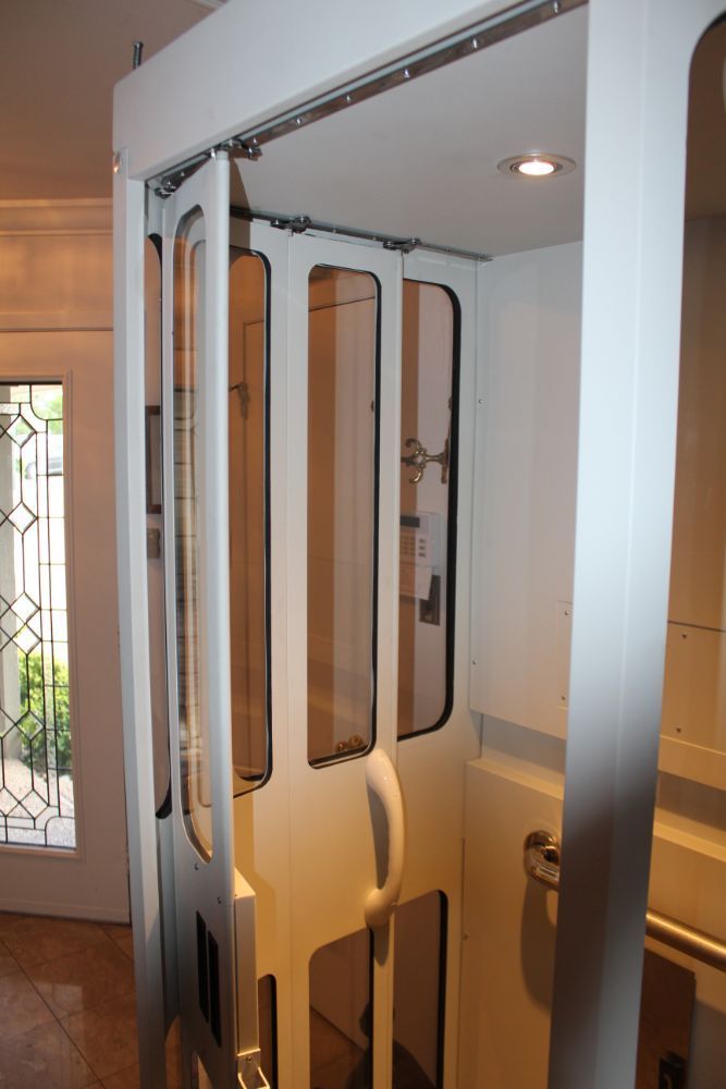 A white residential elevator with sliding, windowed doors standing inside a home near an entryway.
