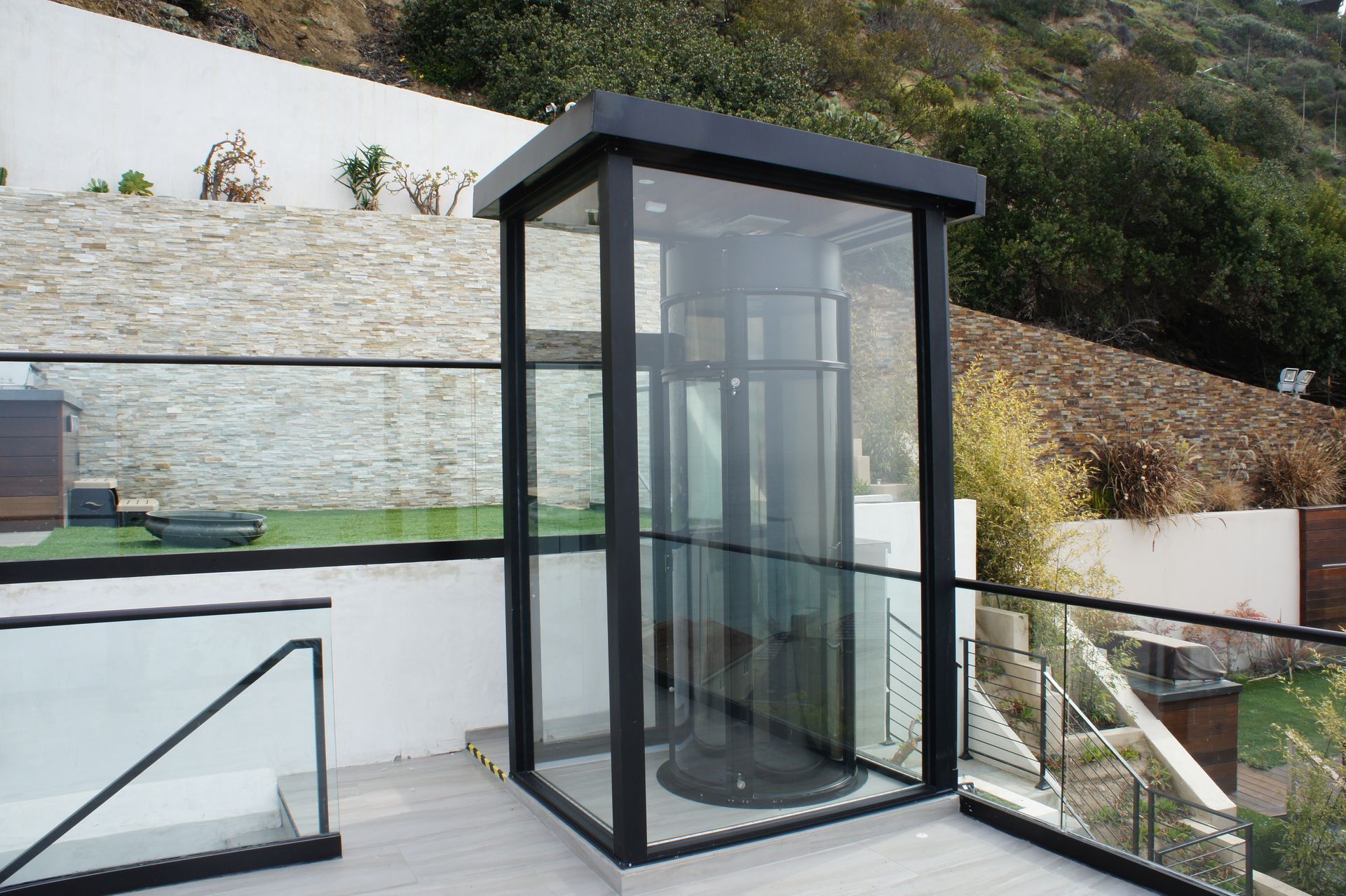 A glass-enclosed outdoor elevator shaft with a black metal frame installed on a modern deck against a hillside.
