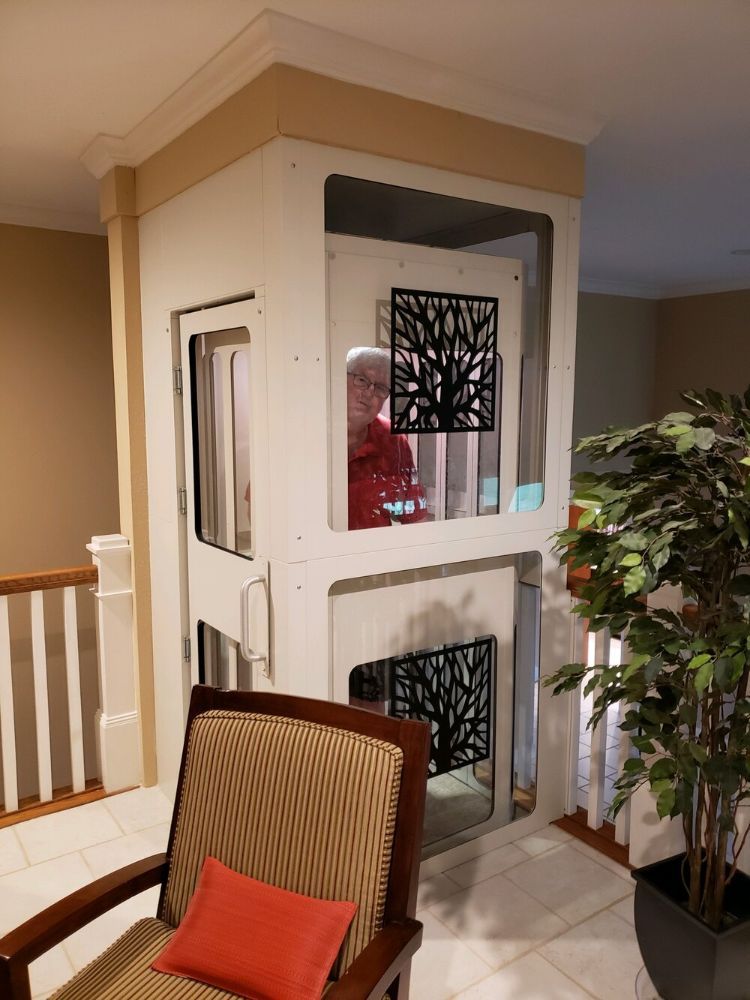A person stands inside a white residential elevator with decorative tree-patterned panels in a carpeted home.