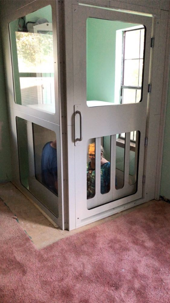 A person sits inside a white residential elevator with large glass panels in a room with pink carpet.