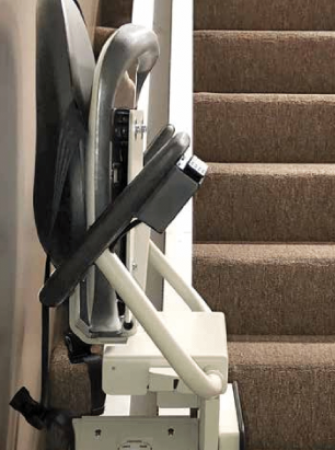 A white home elevator installed in an interior hallway, adjacent to a wooden staircase, a lamp, and a potted plant.