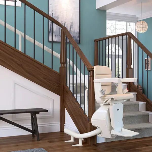 A white stair lift installed on a wooden staircase with teal walls and grey carpeted steps in a residential entryway.