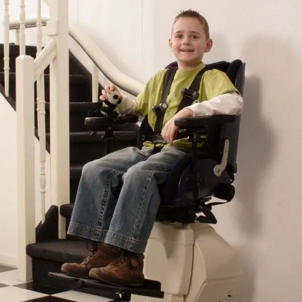 A smiling person seated in a stairlift, wearing a safety harness, positioned at the bottom of a staircase.