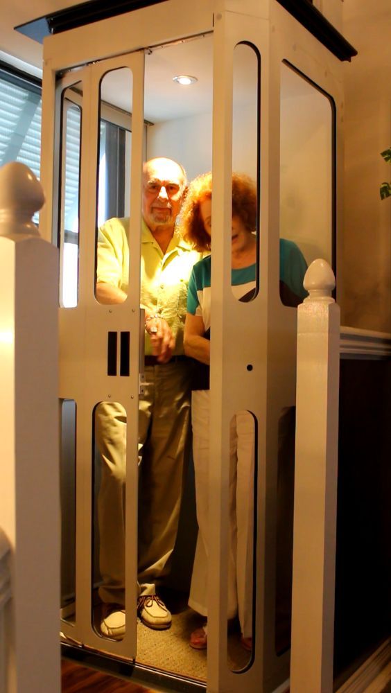 An elderly couple inside a residential glass elevator with white trim.