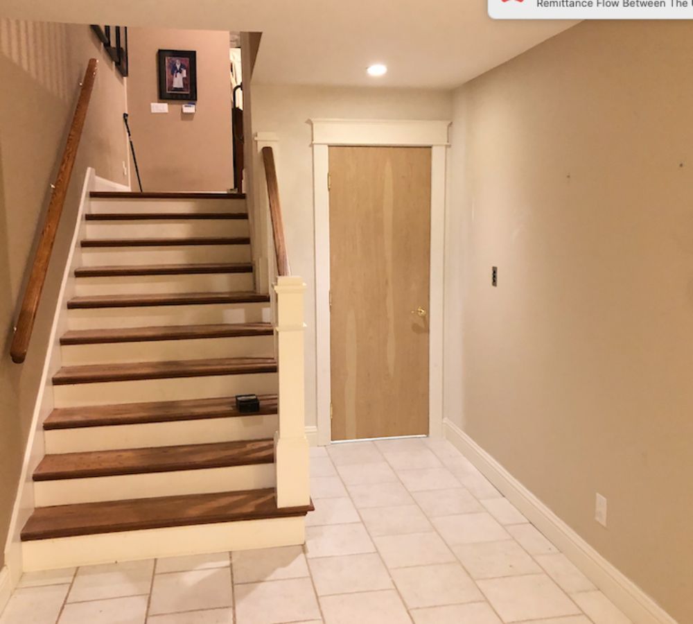 A staircase with dark wood treads and white risers leads upward beside a light-wood door in a tiled foyer.