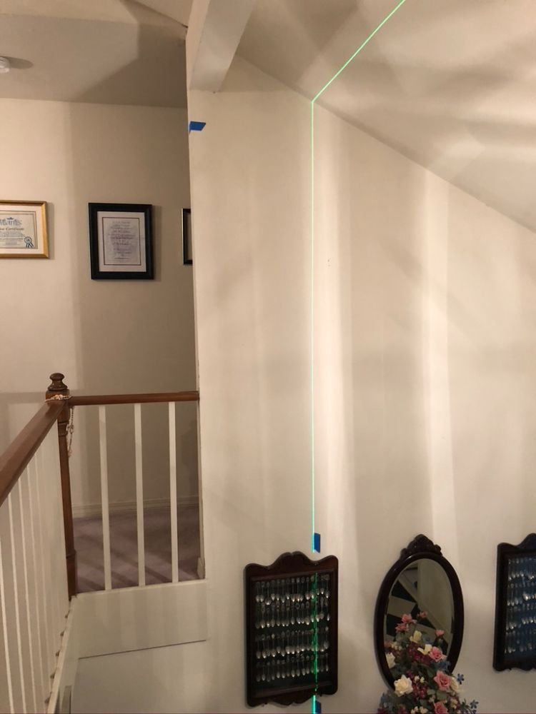 A laser level projects a straight green vertical line down a white interior wall above a stairway and hallway.