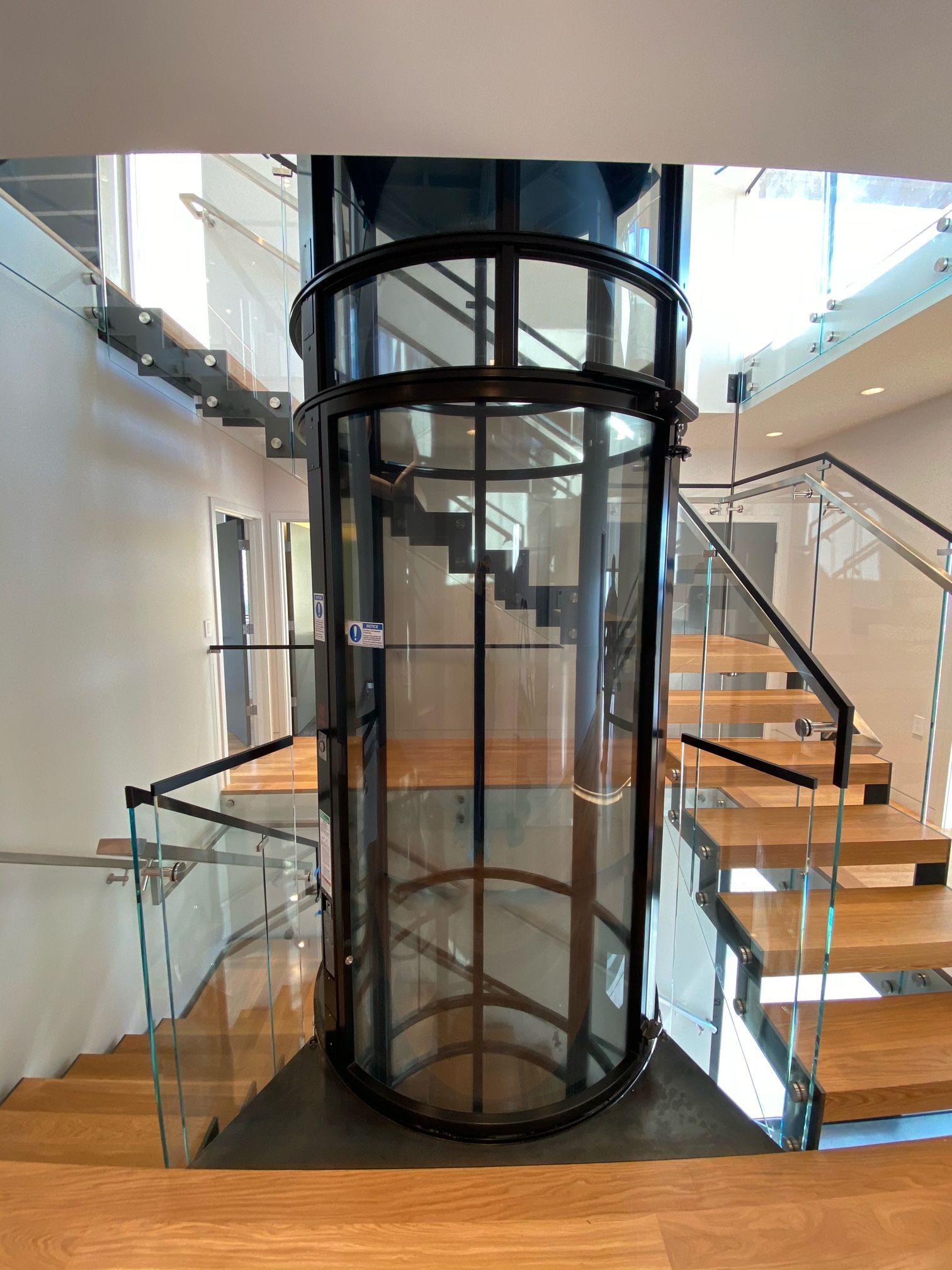 A cylindrical glass elevator stands centrally in a modern house, surrounded by wooden stairs with glass railings.