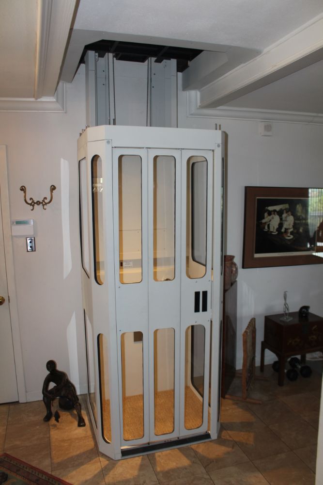 A white residential home elevator with glass panels, shown in a tiled room with a metal support structure above it.