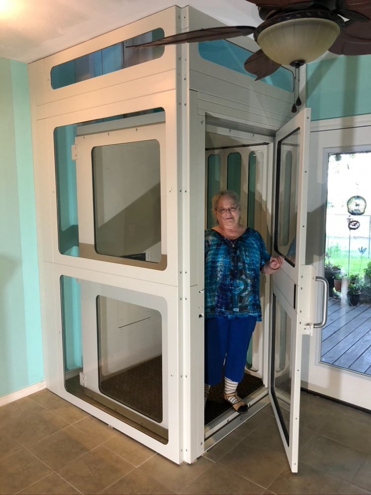 A person stands inside a white, rectangular residential elevator with glass panels in a home setting.