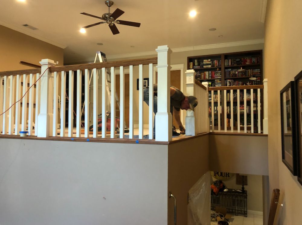 A person installs wooden railing on a landing overlooking an interior space, with a bookshelf visible in the background.