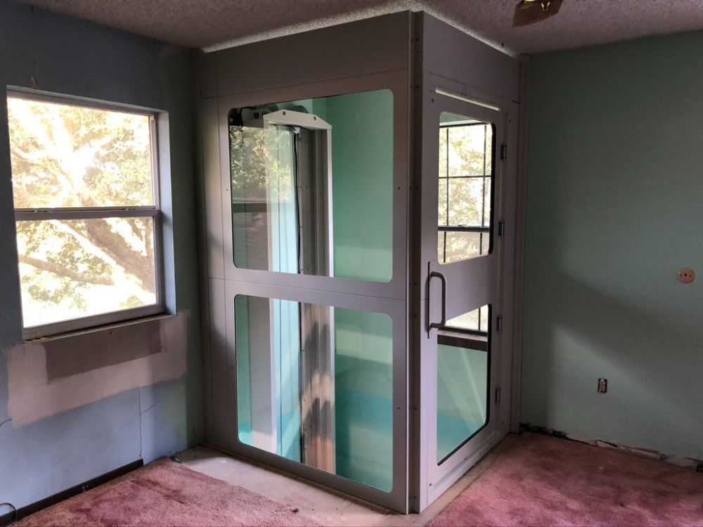 A home elevator with glass panels and a silver metal frame installed in the corner of a room with green walls and carpet.
