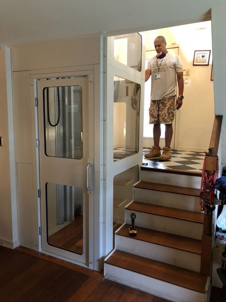 A person stands at the top of a staircase next to a white residential elevator with a glass door in a home interior.