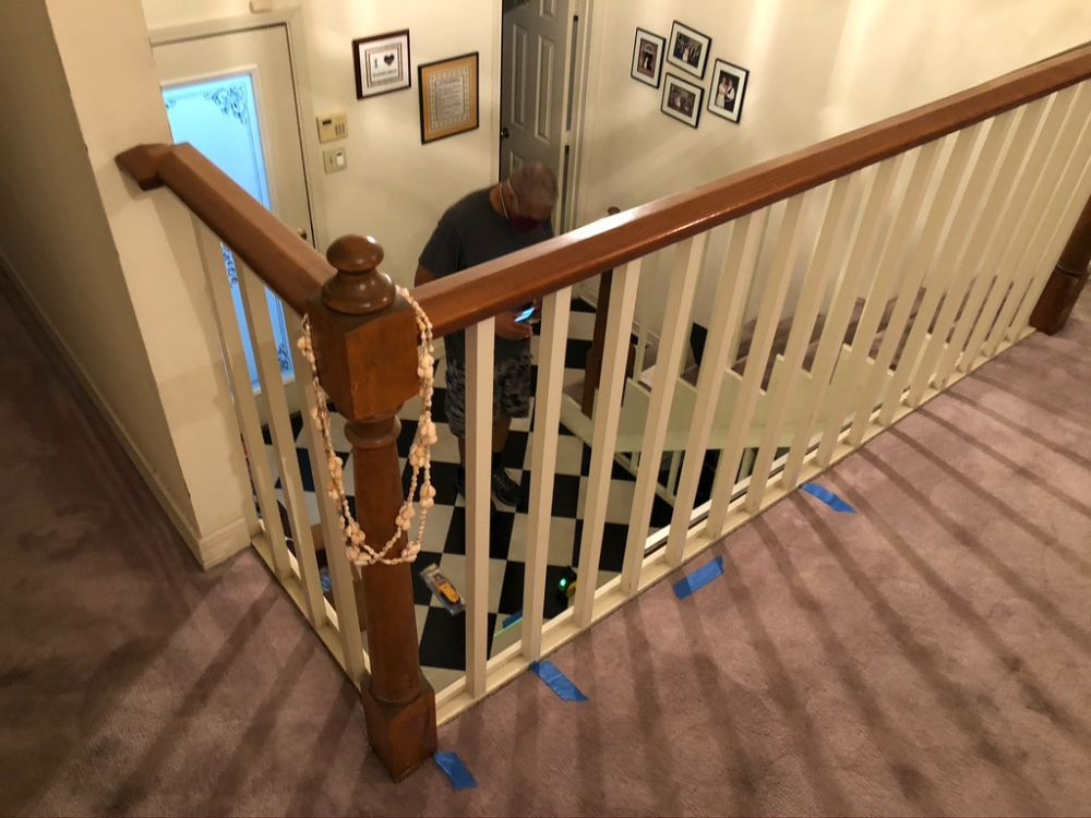 An elevated view of a wooden staircase railing with white spindles, looking down toward a black-and-white tiled entry.
