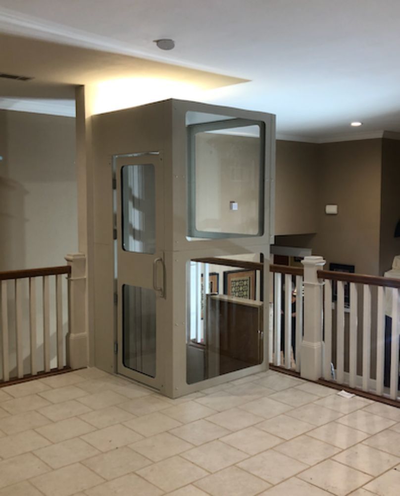 A residential glass-paneled elevator enclosed in a light-colored frame, situated on a tiled landing next to a railing.