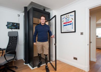 A person with leg prostheses steps out of a small, black home elevator in a bright, modern room.