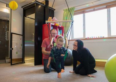 A child sits on a hanging swing in a therapy room, supported by an adult in a red shirt while another adult watches.