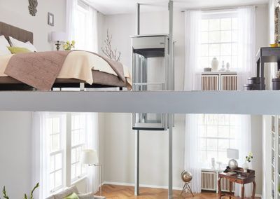 A residential elevator connects a bedroom on an upper loft level to the living space below in a bright, modern home.