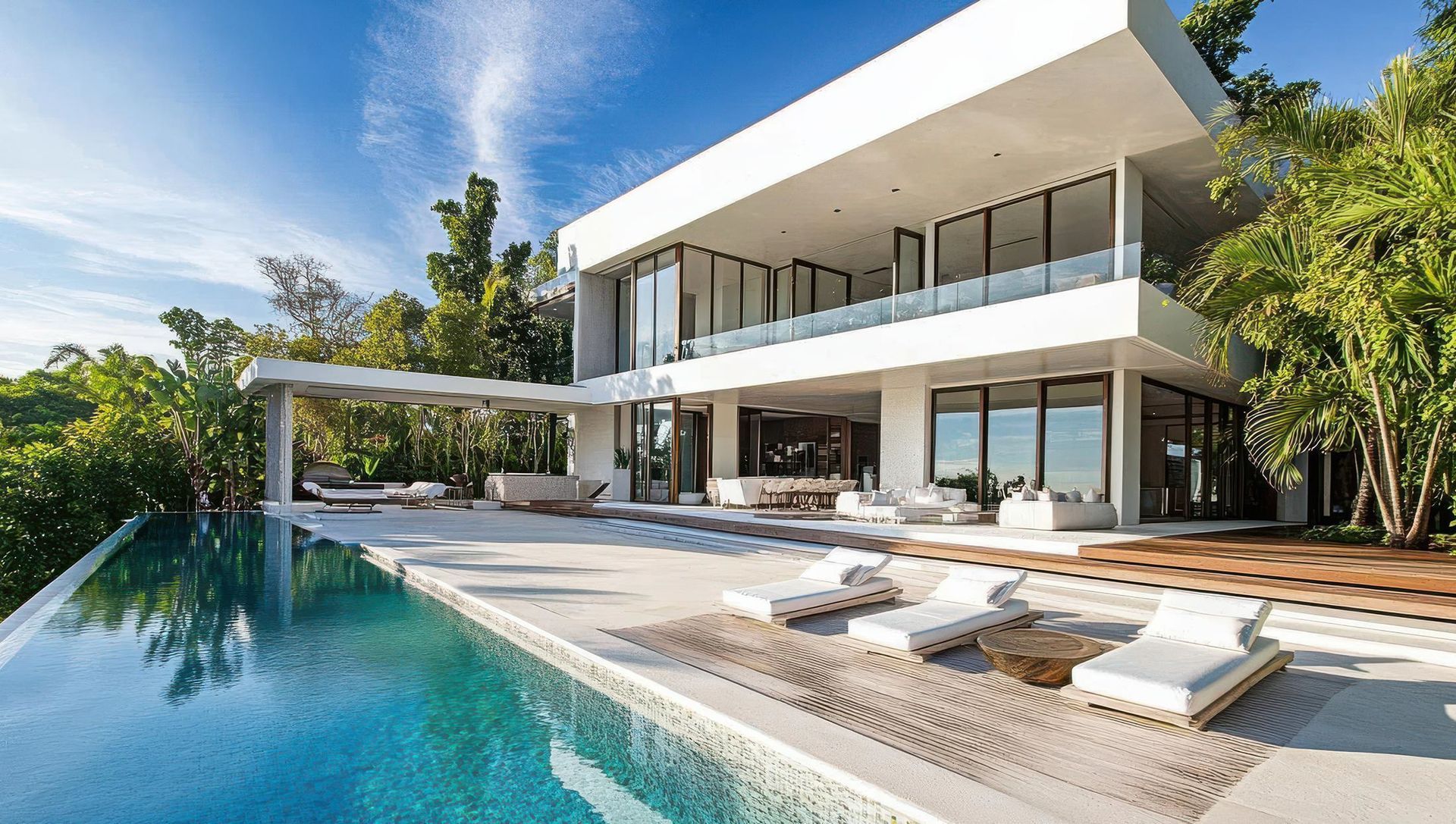 Moderna casa bianca con piscina, sedie a sdraio e vegetazione lussureggiante sotto un cielo azzurro.