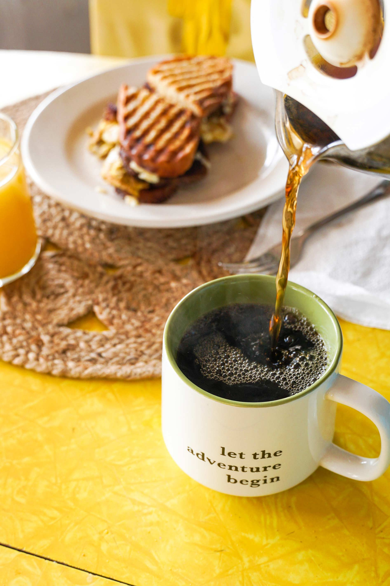 A cup of coffee is being poured from a coffee pot next to a plate of food.
