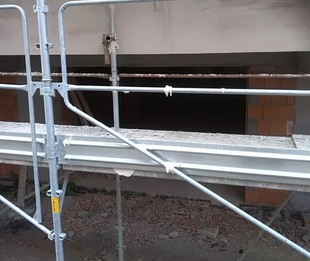 Impalcatura metallica davanti al muro di un edificio, sopra macerie e ghiaia.