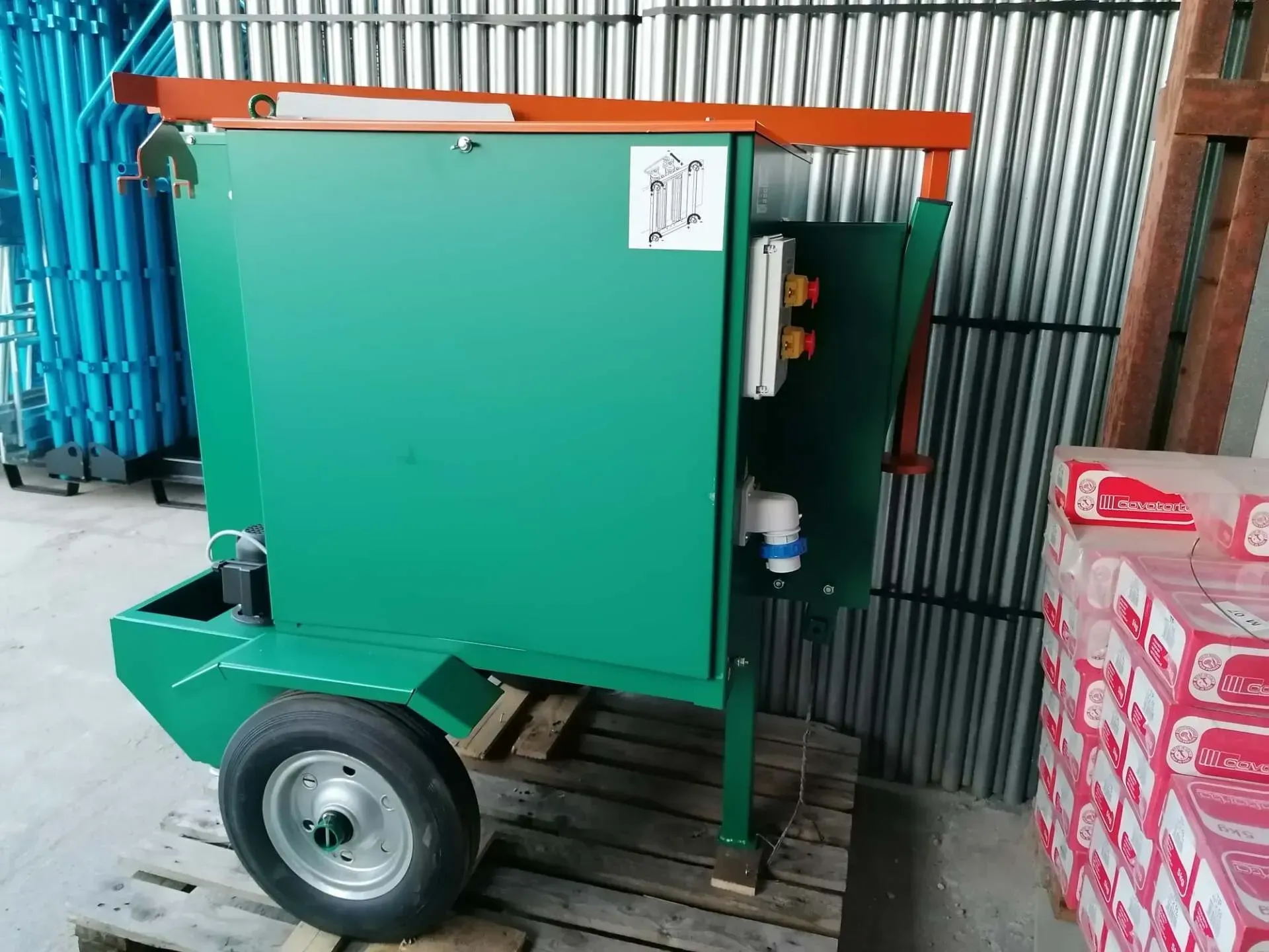 Macchina industriale verde su ruote accanto a una parete di lamiera ondulata in un ambiente di magazzino.