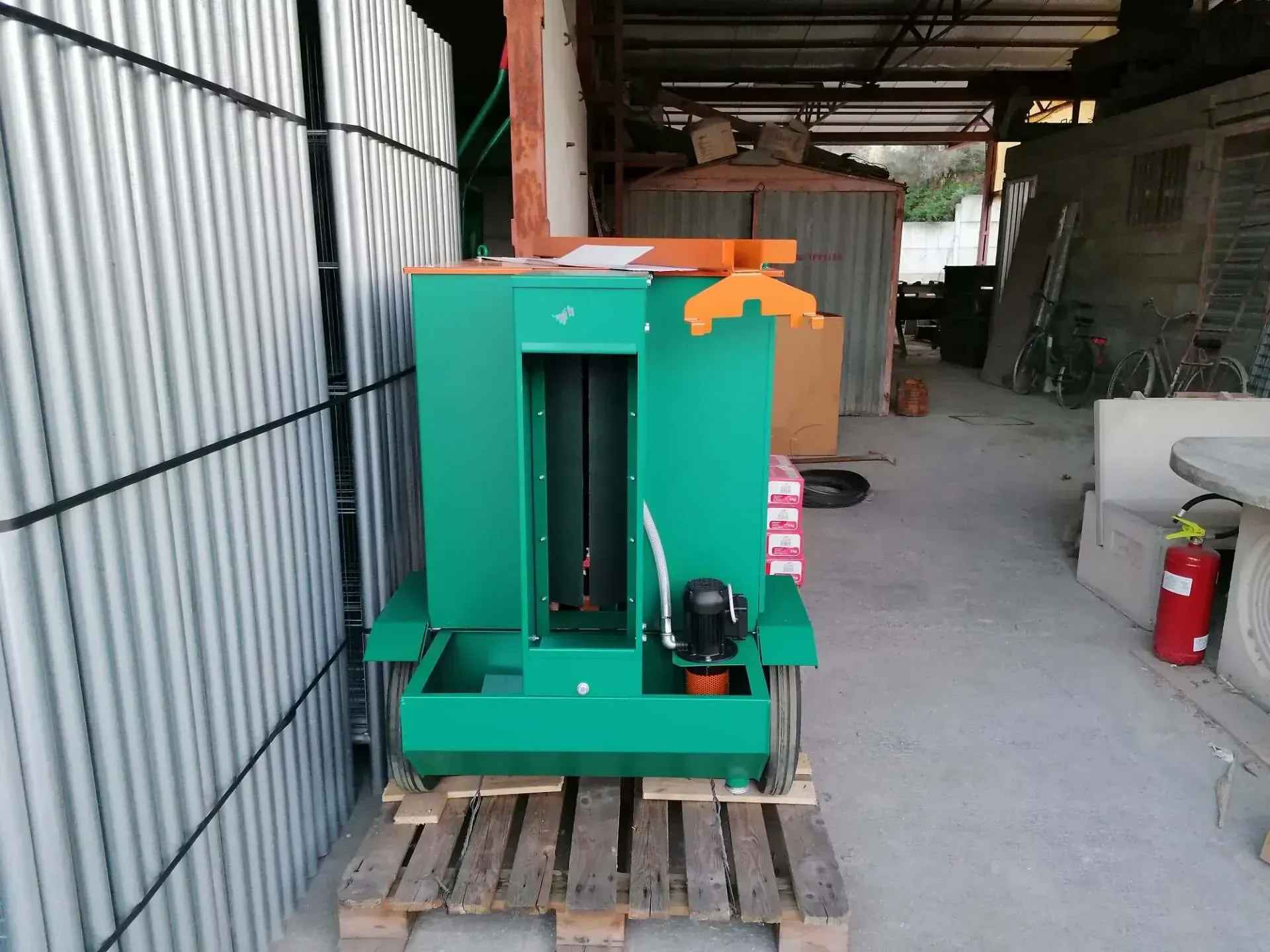 Macchina verde con ruote e tetto arancione all'interno di un capannone in lamiera ondulata, appoggiata su un pallet di legno.