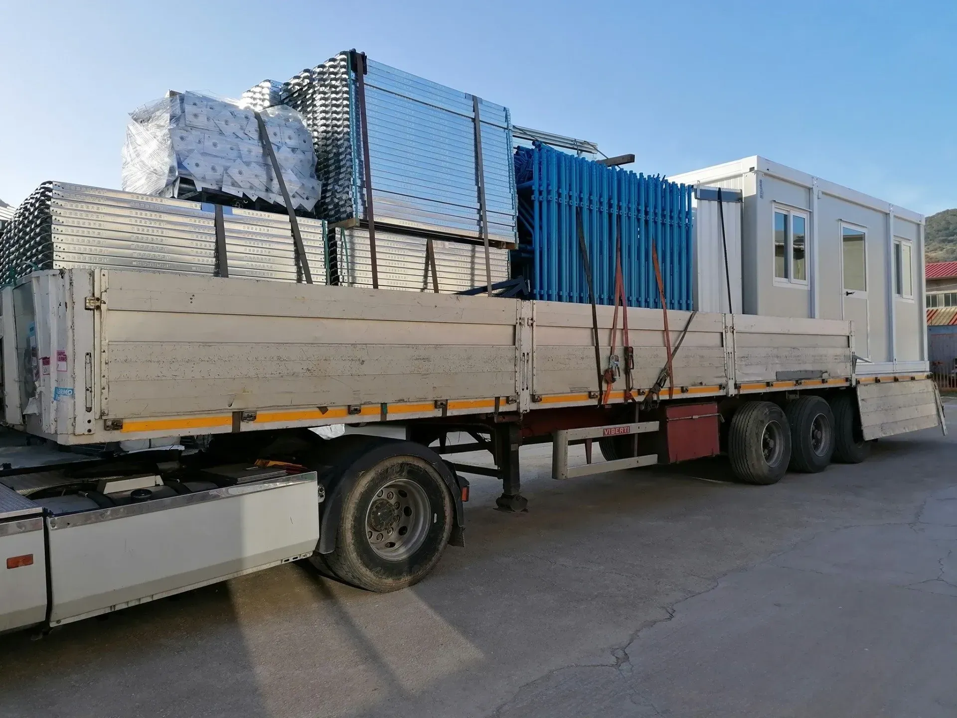 Un camion a pianale ribassato carico di pannelli metallici e materiali imballati in un piazzale industriale.