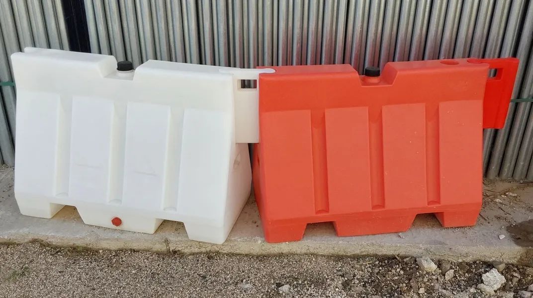 Due barriere stradali in plastica, una bianca e una arancione, appoggiate a un muro di lamiera ondulata.
