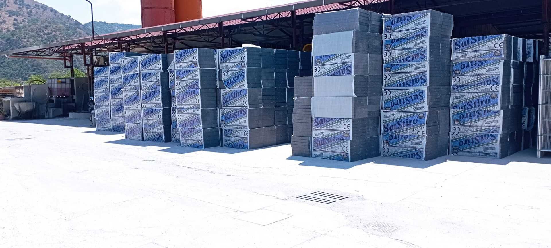 Pile di merci imballate in blu ammassate all'esterno di un magazzino, sotto la luce intensa del sole.
