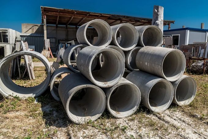 Pile di grandi tubi di drenaggio in cemento in un cortile accanto a un capannone e a un edificio blu.