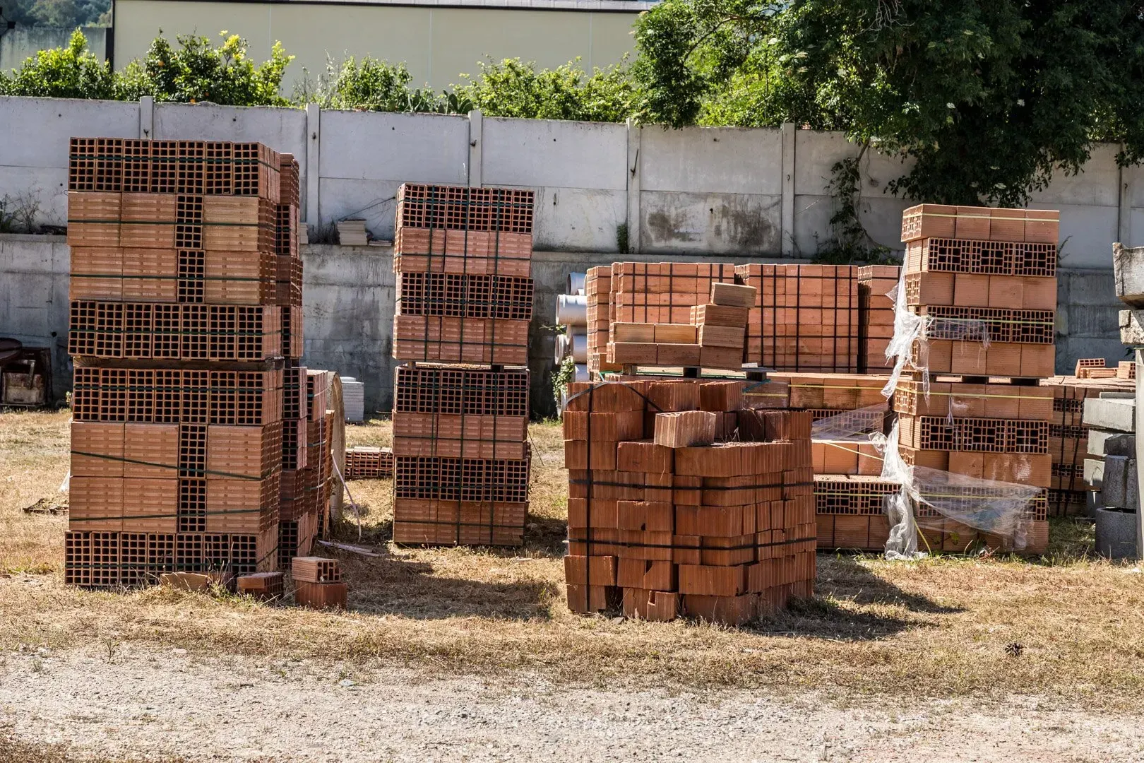 Pile di mattoni di argilla rossa accatastate all'aperto su terreno asciutto accanto a un muro di cemento.