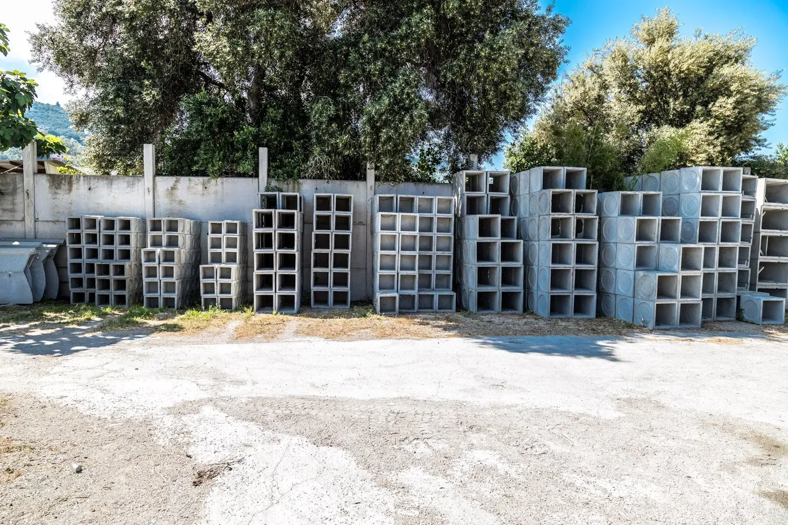 Pile di blocchi e lastre di cemento grigio per il drenaggio su un piazzale ghiaioso, con grandi tubi di cemento sullo sfondo.