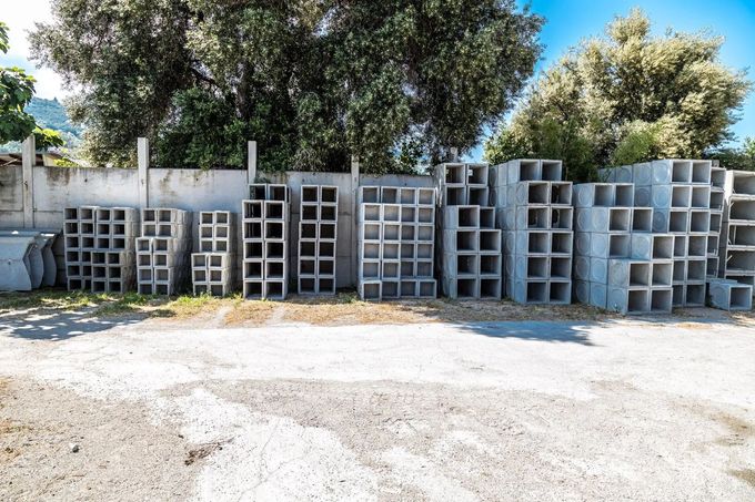 Pile di blocchi di cemento grigio allineate all'aperto, sotto gli alberi, a lato di una strada sterrata.