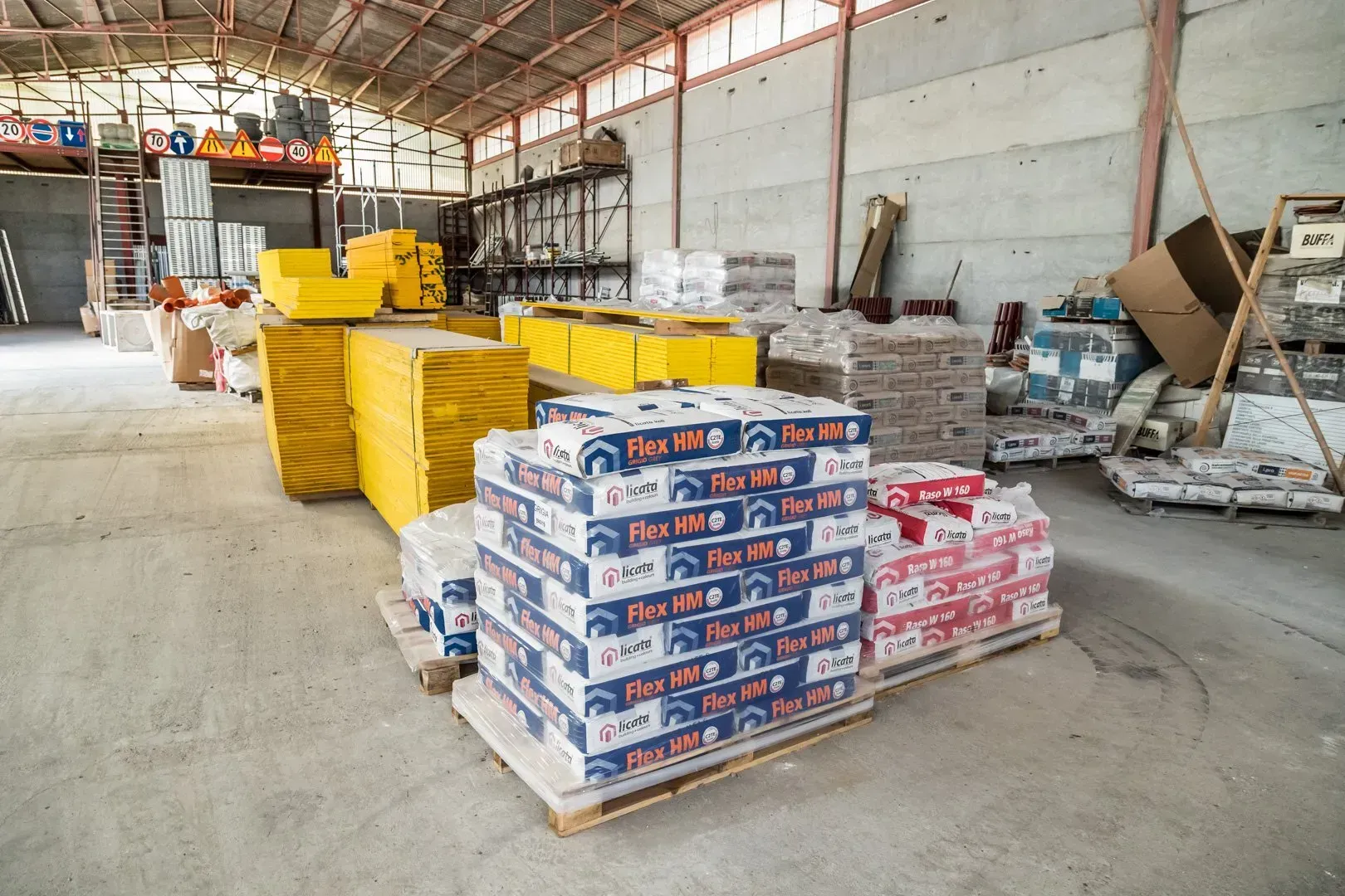 Pallet di materiali edili impilati in un magazzino, con cartelli di avvertimento sulla parete di fondo.