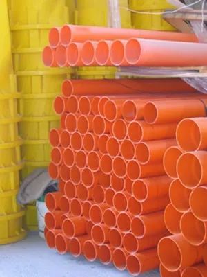Pile di grossi tubi neri con estremità arancioni, stoccati all'aperto accanto a un edificio industriale.