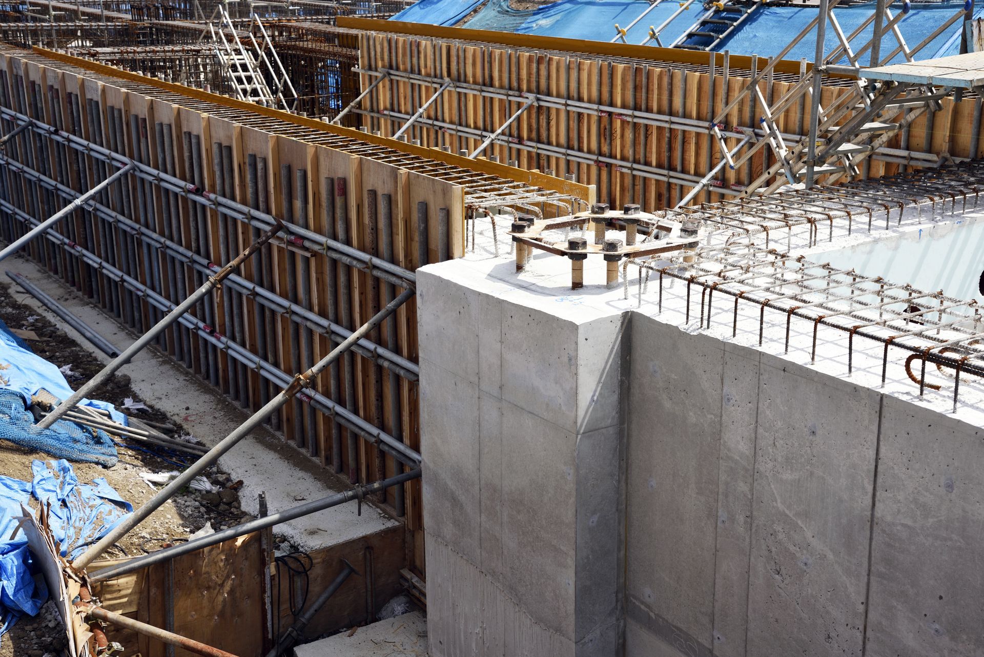 Armatures et coffrage en bois autour d'une fondation en béton en construction, avec des échafaudages et une bâche bleue à proximité.