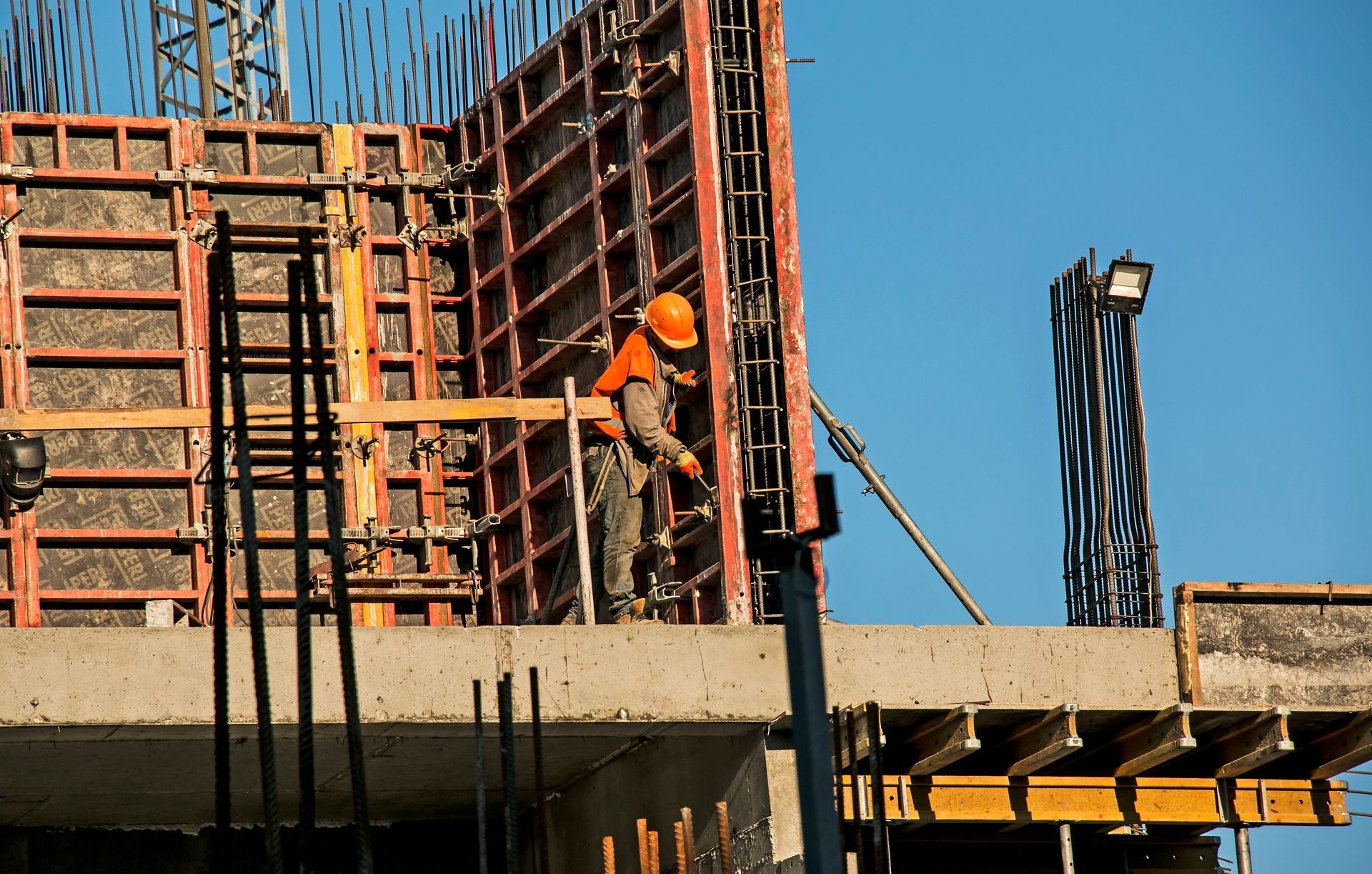 Ouvrier du bâtiment portant un casque orange sur un échafaudage, à côté d'une dalle de béton et d'armatures métalliques, sous un ciel bleu.