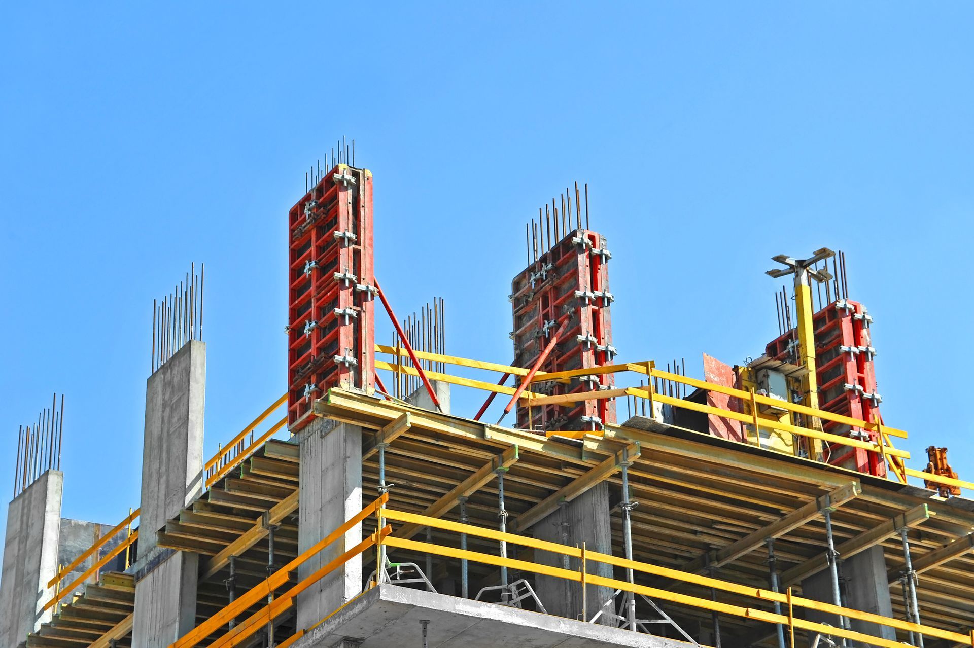 Chantier de construction avec des colonnes en béton, des échafaudages rouges et des rambardes jaunes sur fond de ciel bleu