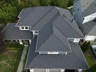 Aerial view of a house with a dark gray shingled roof and white trim in a wooded neighborhood