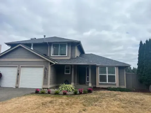 Two-story gray house with white garage doors, front porch, and small flower bed under a cloudy sky